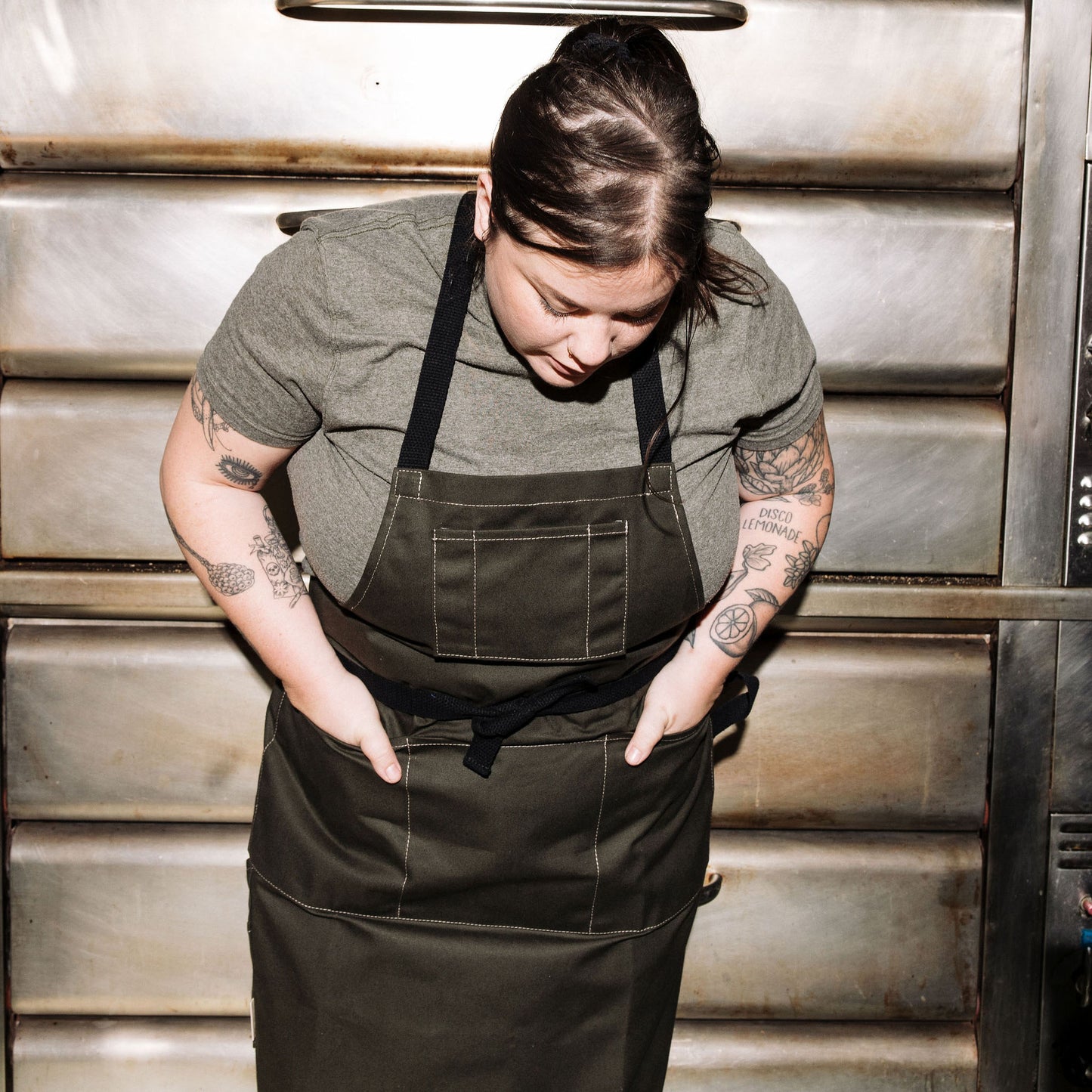 Person wearing a pine canvas apron standing in front of stainless steel oven doors in a commercial kitchen