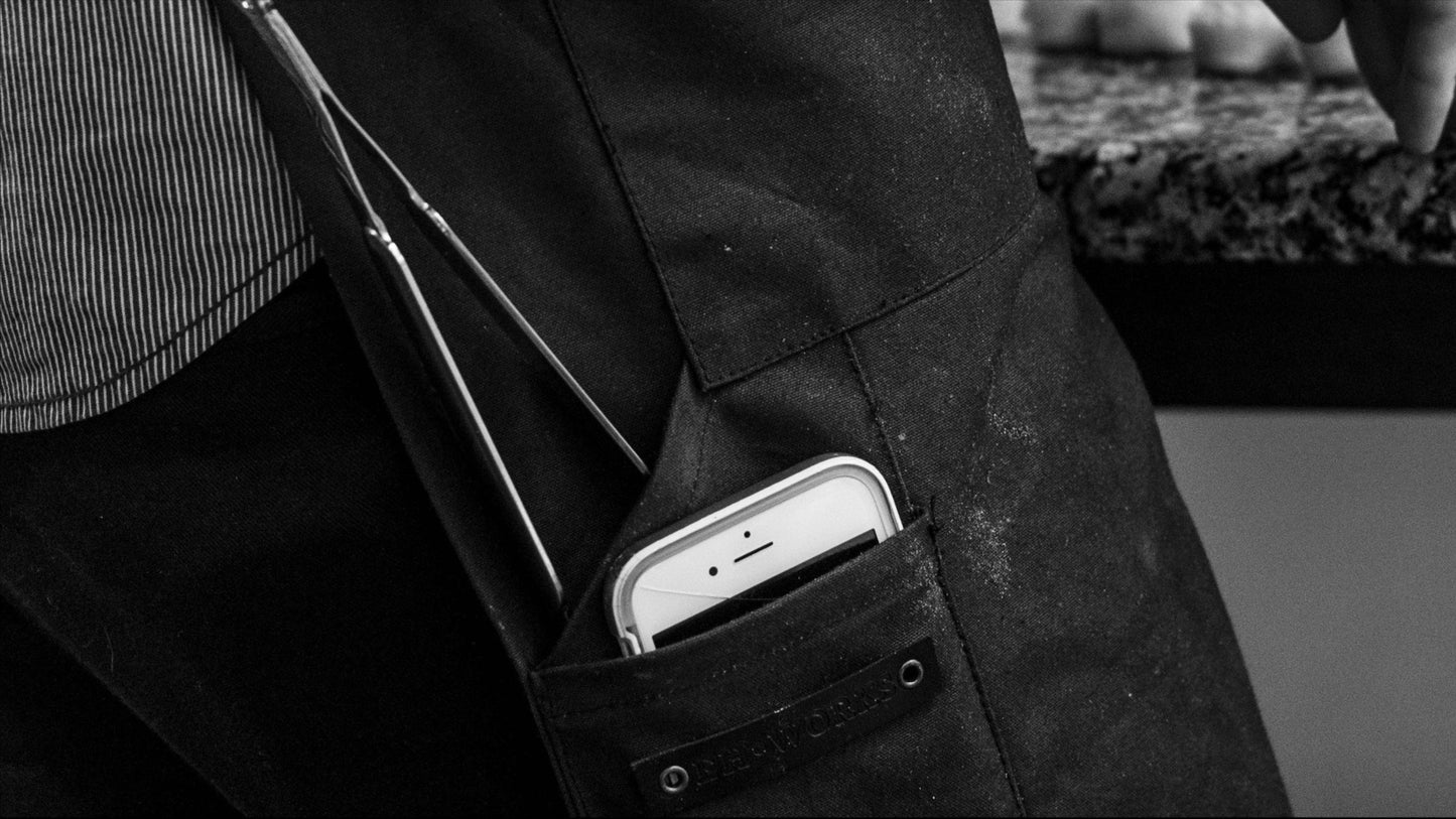 Black-and-white close-up of a pine canvas apron pocket holding a smartphone and tweezers, EH·Works label visible