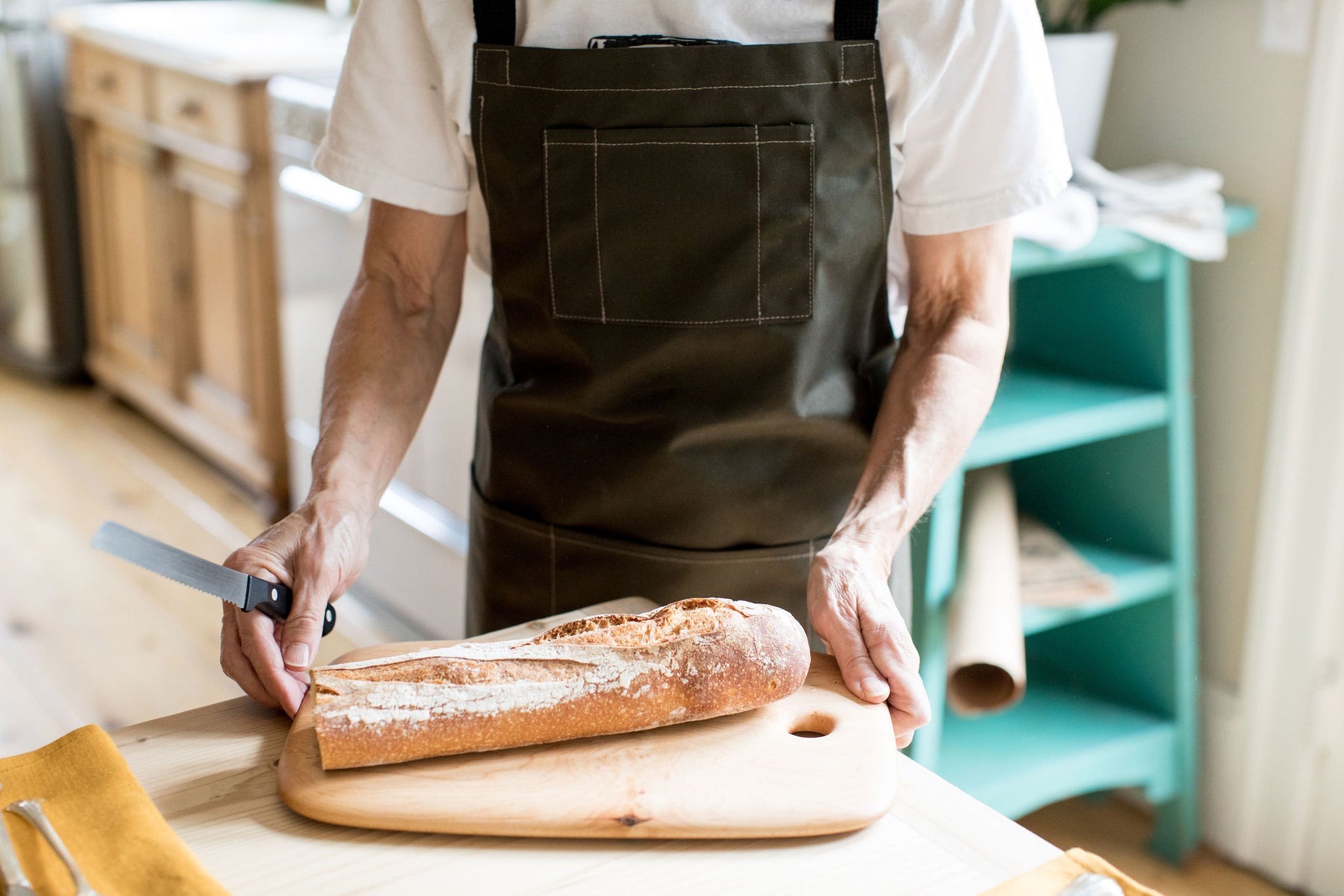 Person wearing a pine canvas apron while slicing a baguette on a wooden cutting board in a kitchen