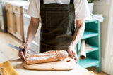 Person wearing a pine canvas apron while slicing a baguette on a wooden cutting board in a kitchen