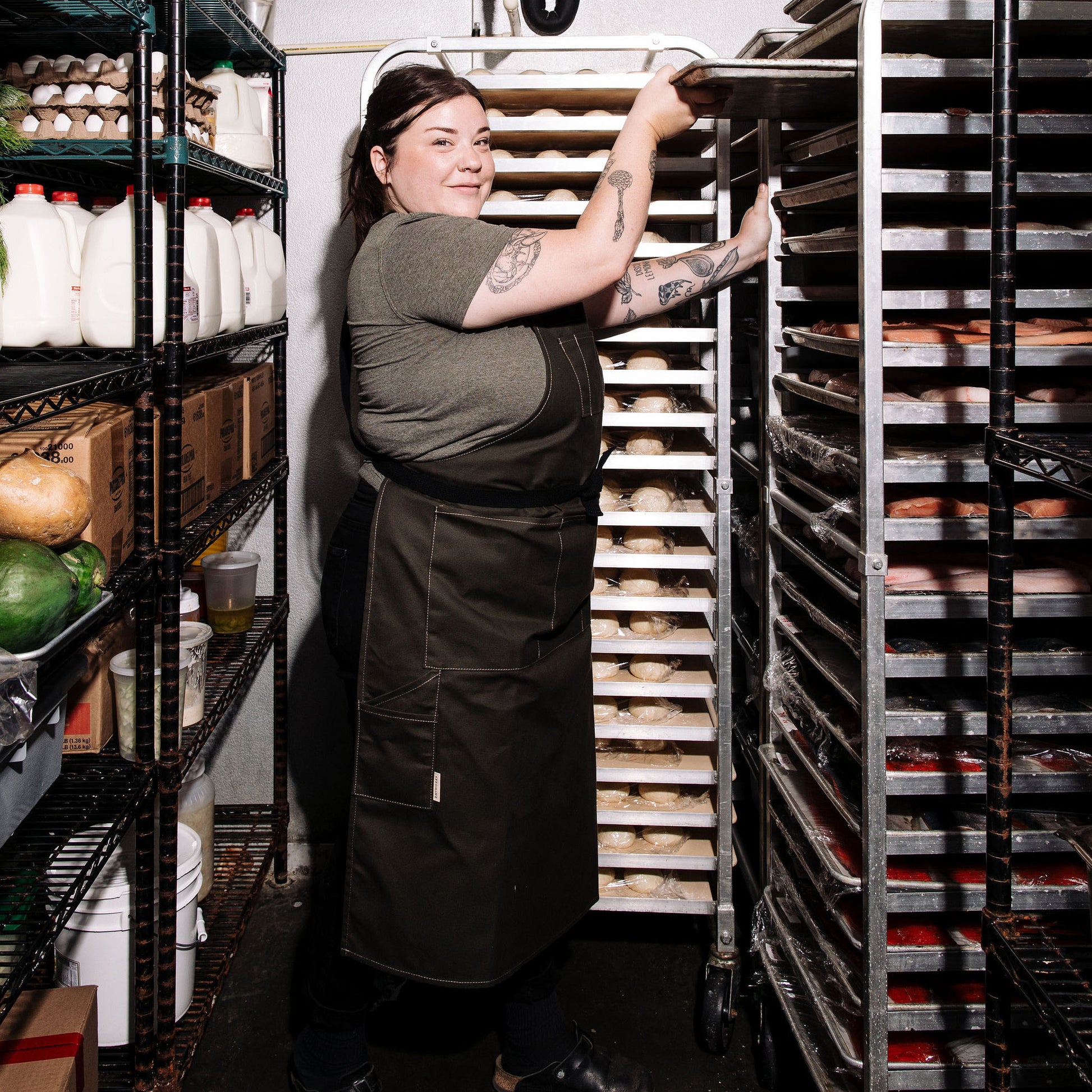  Person wearing a pine canvas apron while sliding a baking tray into a rack in a walk-in cooler