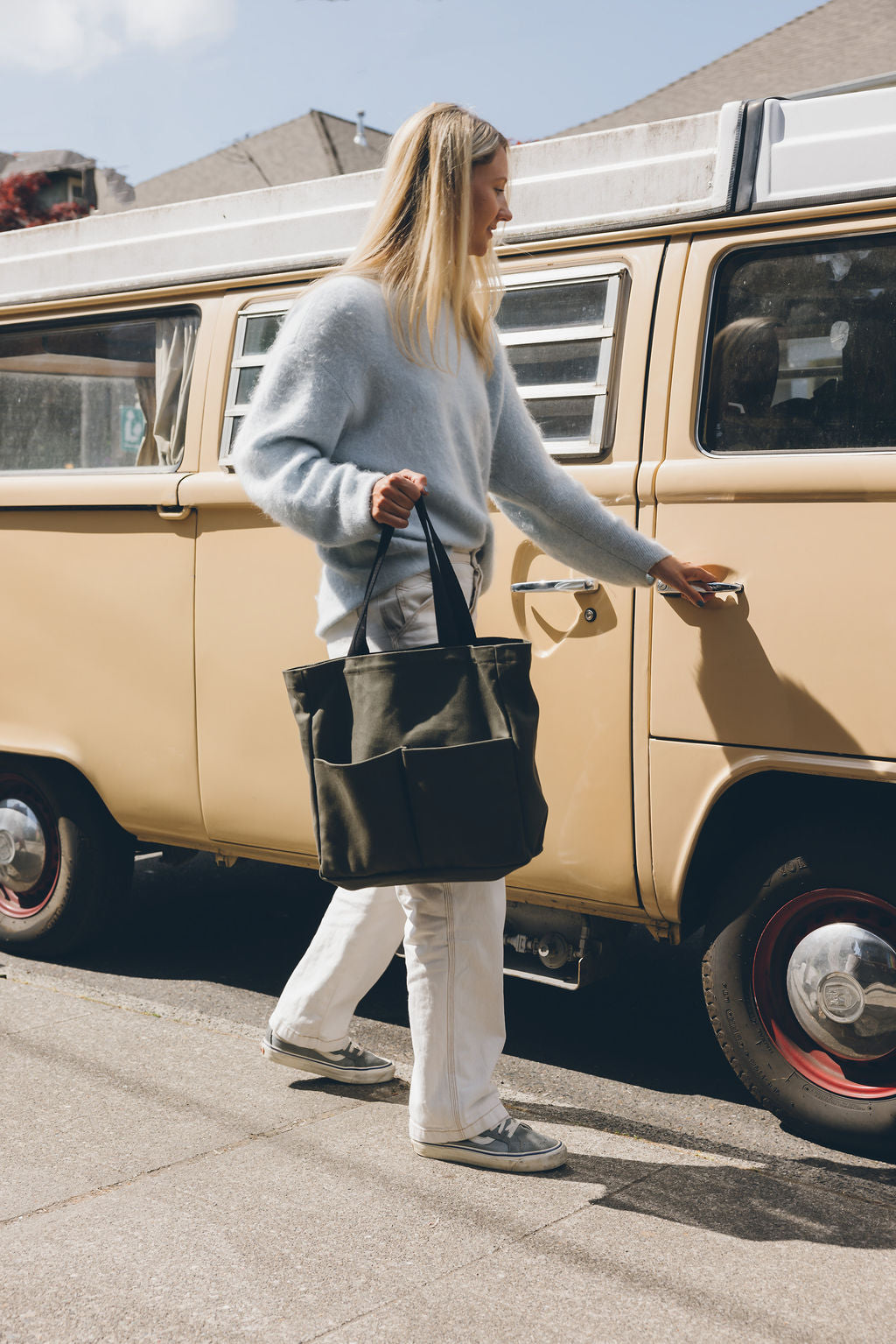 Woman carrying an olive green canvas tote bag with front pockets and black handles while walking beside a vintage van
