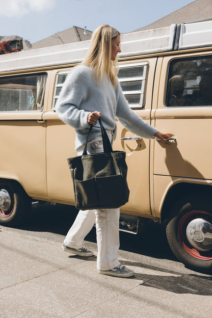 Woman carrying an olive green canvas tote bag with front pockets and black handles while walking beside a vintage van
