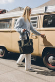 Woman carrying an olive green canvas tote bag with front pockets and black handles while walking beside a vintage van

