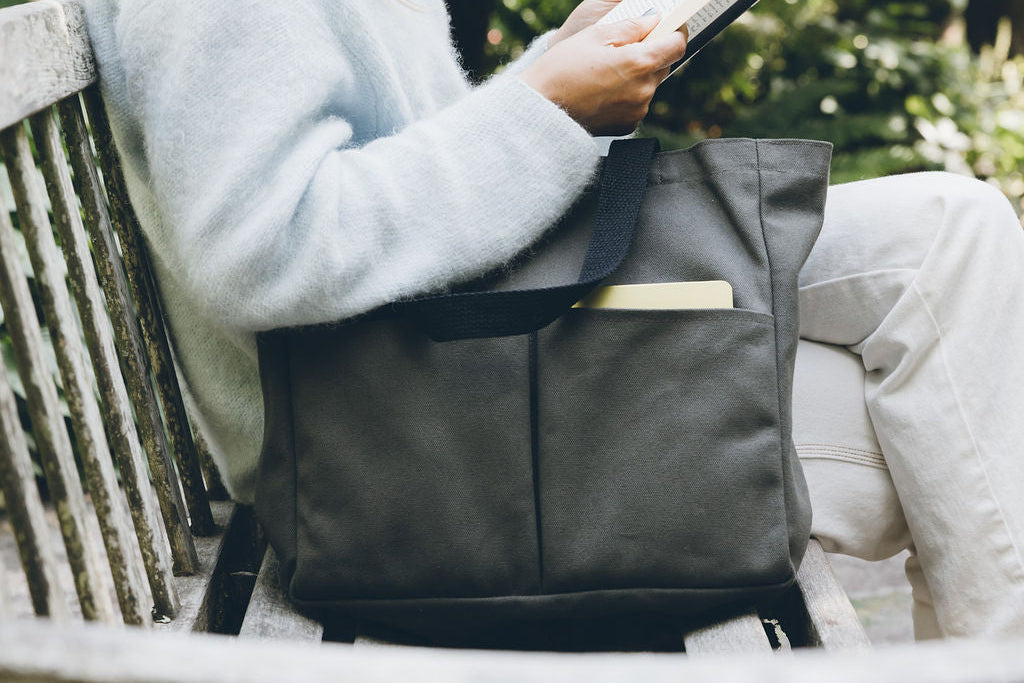 Olive green canvas tote bag resting on a bench beside a woman reading, with a notebook visible in the side pocket
