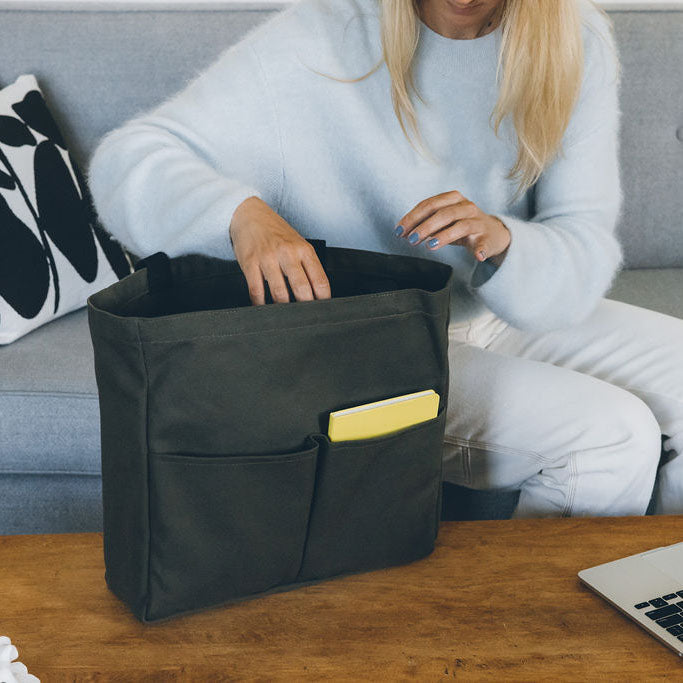 Woman reaching into an olive green canvas tote bag with front pockets, showing the interior while sitting near a laptop
