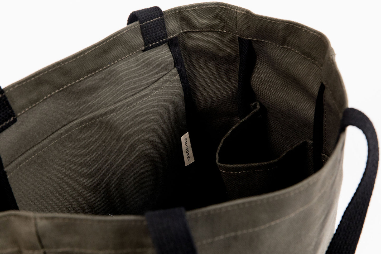 Close-up of an olive green canvas tote bag interior, showing the EH·Works brand label and stitched pocket construction
