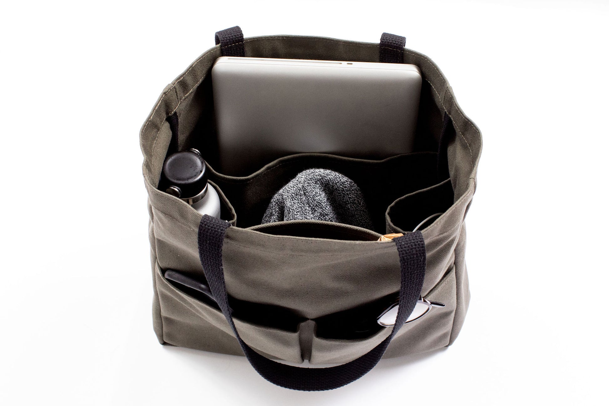 Olive green canvas tote bag open and packed with a laptop, water bottle, sweater, sunglasses, and pen, showing interior organizer pockets
