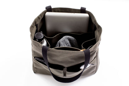 Olive green canvas tote bag open and packed with a laptop, water bottle, sweater, sunglasses, and pen, showing interior organizer pockets
