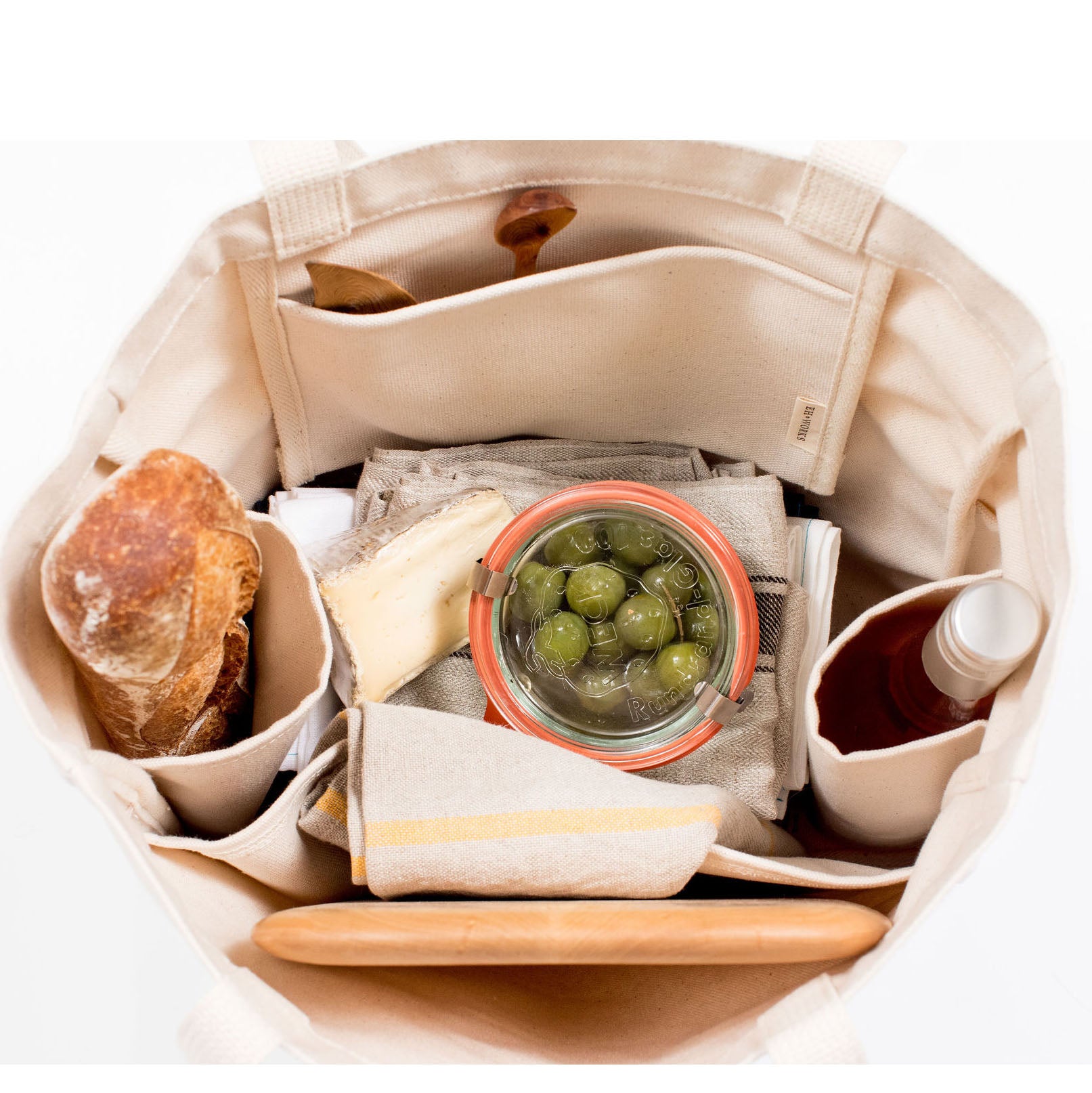 Open natural canvas Utility Carryall tote packed with bread, olives, cheese, and a bottle on a white background