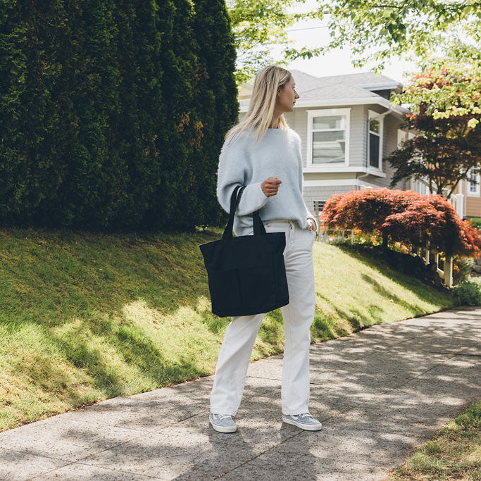 Woman carrying the Canvas Utility Carryall tote in black, walking on a sidewalk with a house and garden in the background