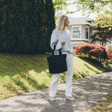 Woman carrying the Canvas Utility Carryall tote in black, walking on a sidewalk with a house and garden in the background