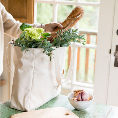 Person holding a baguette over a natural canvas Utility Carryall tote filled with groceries in a kitchen