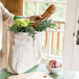 Person holding a baguette over a natural canvas Utility Carryall tote filled with groceries in a kitchen