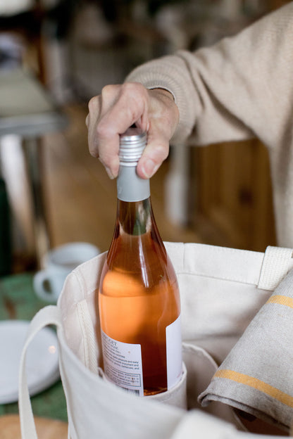 Person placing a bottle of rosé wine into a natural canvas Utility Carryall tote in a casual outdoor setting