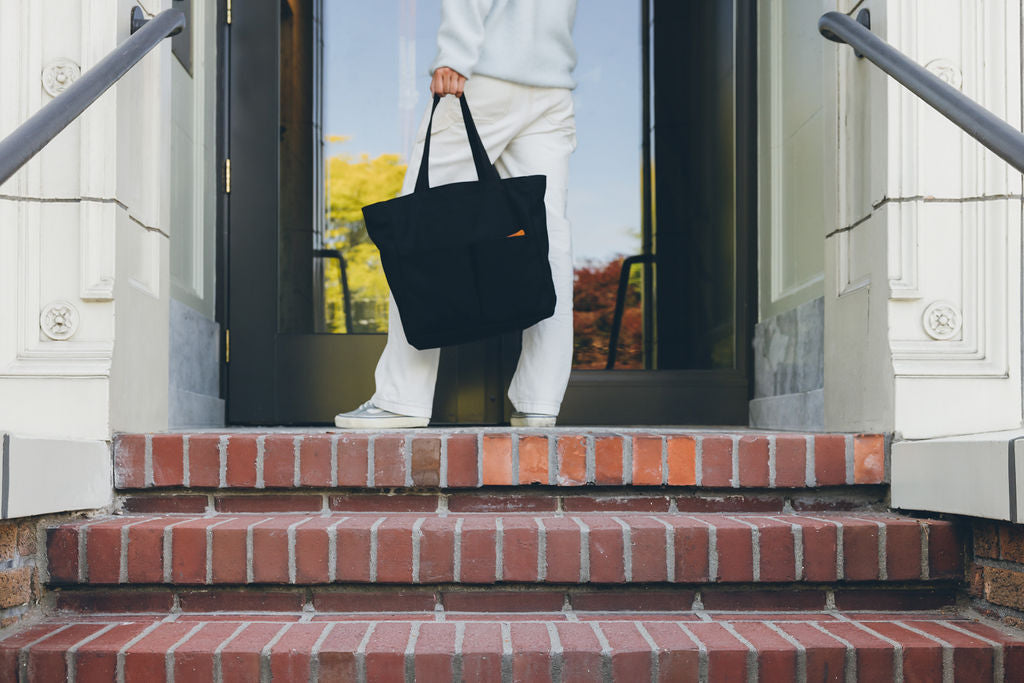 Person holding the Canvas Utility Carryall tote in black by its handles, stepping down brick steps from a doorway