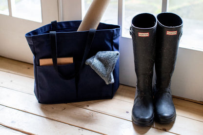 Canvas Utility Carryall tote in navy, holding a rolled poster and beanie, next to black rain boots by a rustic window entryway