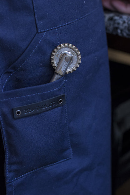 Close-up of a navy canvas apron pocket with a pasta cutting tool and the EH·Works leather brand label