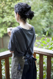 Person wearing an army green canvas apron with crossback straps while holding a cup on an outdoor balcony