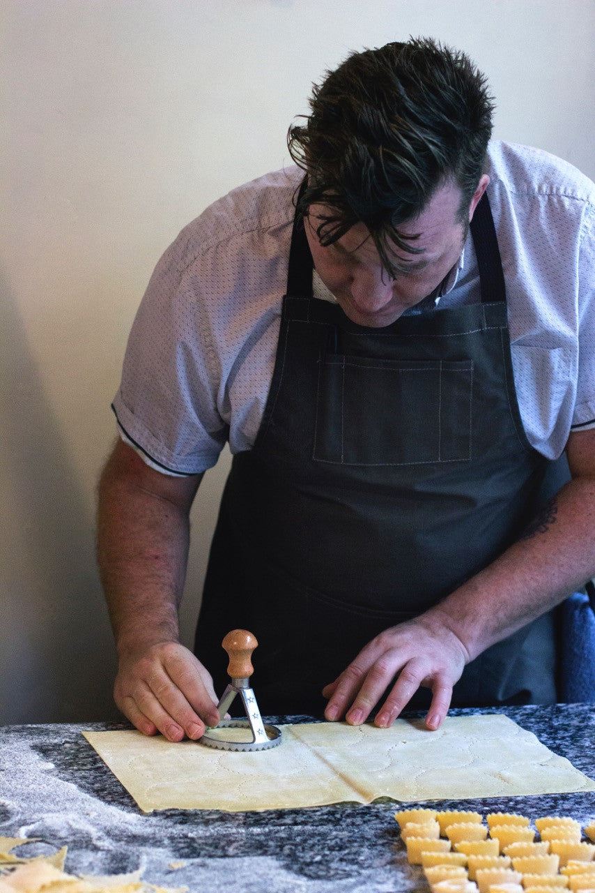 Person wearing a pine canvas apron while cutting fresh pasta dough on a floured countertop