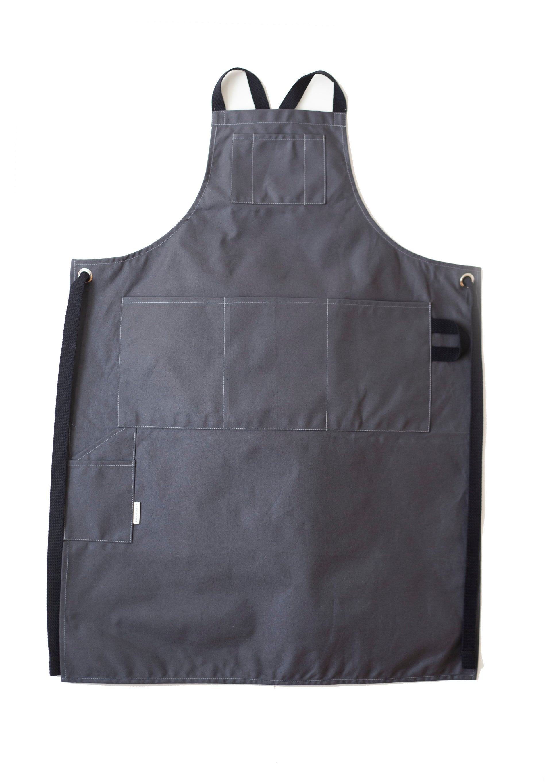 Grey canvas apron with multiple pockets and adjustable crossback straps, laid flat on a white background
